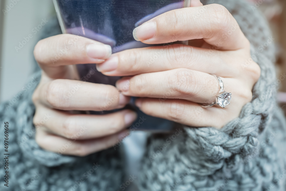 Close up of an elegant engagement diamond ring on woman finger with dark gray sweater winter clothe while holding mobile phone . love and wedding concept. Soft and selective focus.