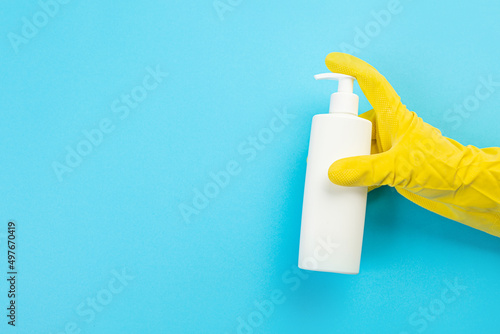 A hand in a yellow glove holds a white bottle of detergent on a blue background. Banner with copy space. Chemical cleaners, household chemicals, brushes and collage supplies. Cleaning concept.