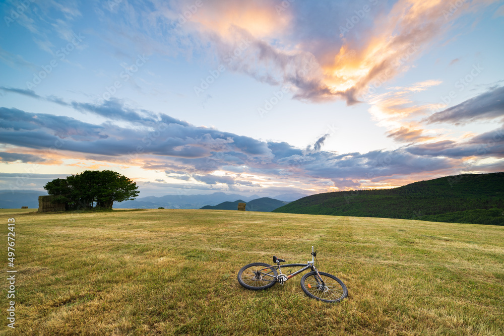 Sunset dramatic sky over meadow in Marche region, Italy. MTB lying on grass amid unique hills and mountains landscape. Traveling by bicycle, alternative mobility concept.