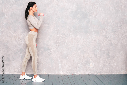 Fitness confident woman in sports clothing. Sexy young beautiful model with perfect body. Female posing near grey wall in studio. Stretching out before training.Making exercises with dumbbells