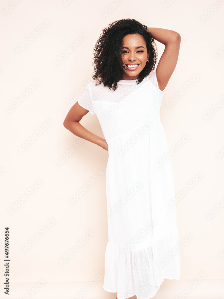 Beautiful black woman with afro curls hairstyle. Smiling model dressed in white summer dress. Sexy carefree female posing near wall in studio. Tanned and cheerful. Isolated Beautiful black woman with afro curls hairstyle. Smiling model dressed in white summer dress. Sexy carefree female posing near wall in studio. Tanned and cheerful. Isolated