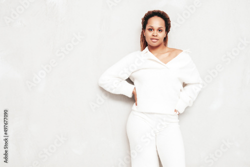 Beautiful black woman with afro curls hairstyle. Smiling model dressed in white summer costume. Sexy carefree female posing near grey wall in studio. Tanned and cheerful. Isolated