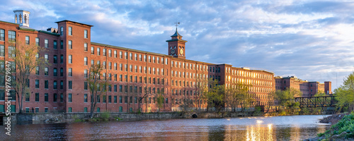 Historic cotton mill building with clock tower in an old industrial park on the Nashua River illuminated by the sun during sunset in May. Panoramic photography. Nashua, New Hampshire, USA