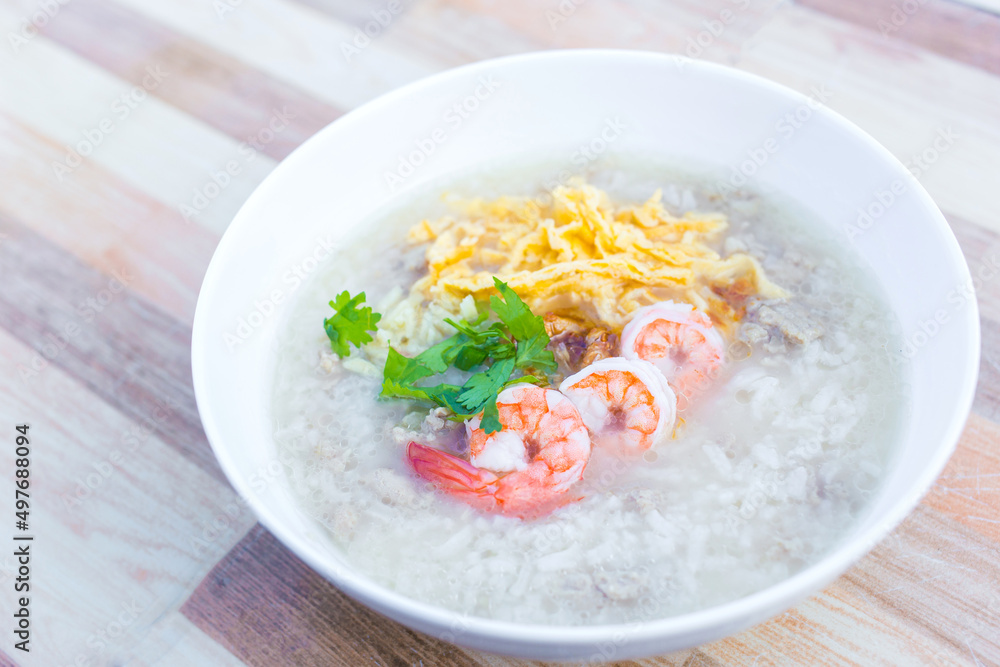 Boiled rice with pork was added on top with chopped omelet, boiled shrimp and coriander as a a breakfast meal in the morning in Thai style while that bowl put on wooden background. Boiled rice with pork was added on top with chopped omelet, boiled shrimp and coriander as a a breakfast meal in the morning in Thai style while that bowl put on wooden background.