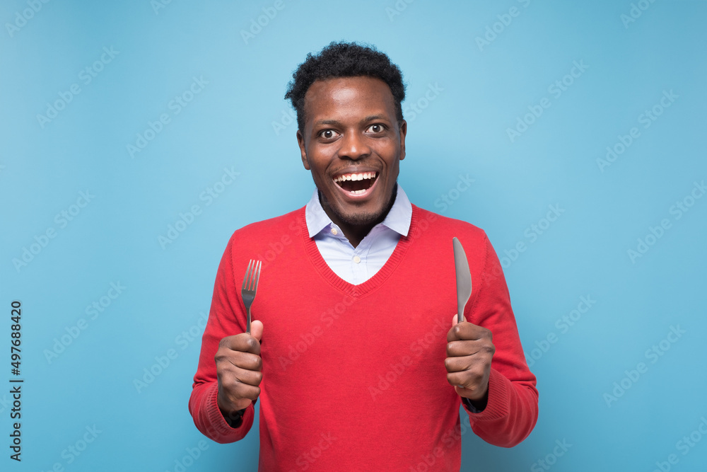 Hungry african young man holding fork and knife on hand ready to eat Hungry african young man holding fork and knife on hand ready to eat