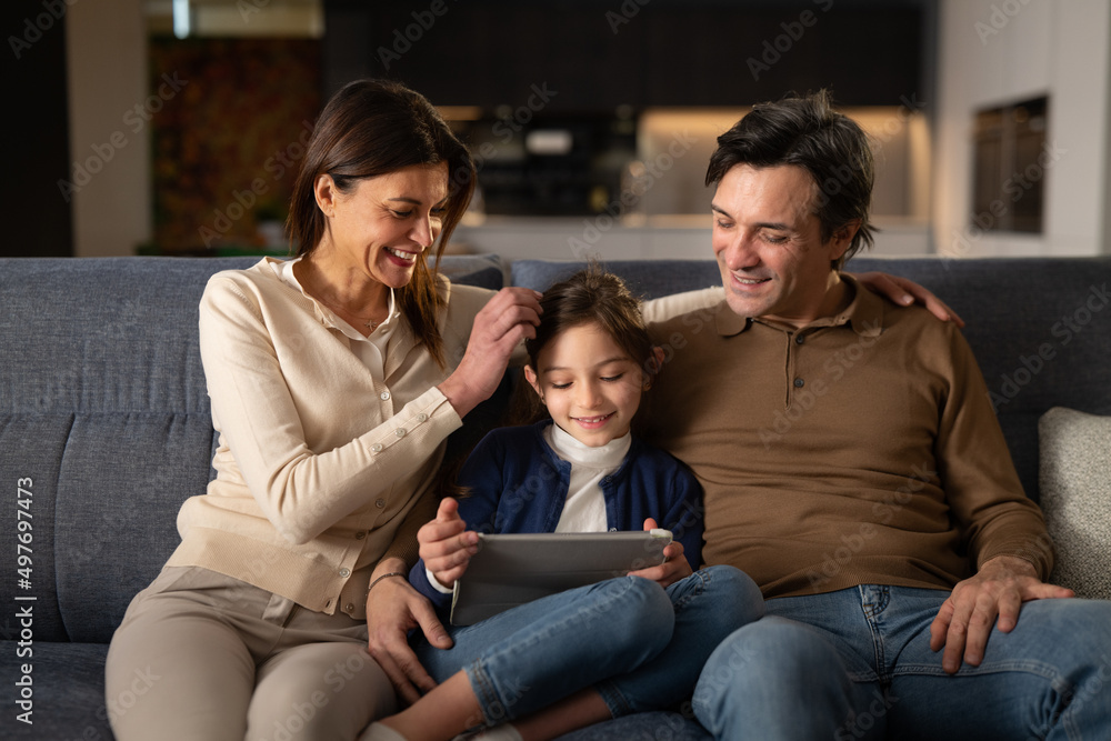Cinematic shot of happy young parents and their cute little girl daughter is smiling in camera while using technology digital tablet together for family entertainment on sofa in living room at home. Cinematic shot of happy young parents and their cute little girl daughter is smiling in camera while using technology digital tablet together for family entertainment on sofa in living room at home.