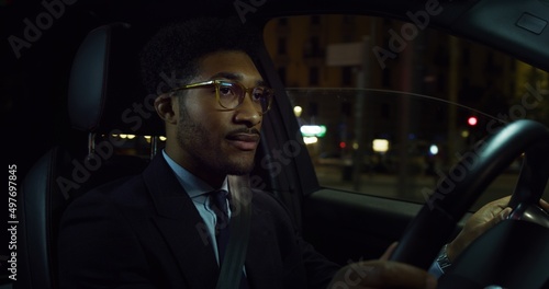 Cinematic shot of young handsome afro american successful elegantly dressed businessman with glasses is driving modern luxury car in center of the city by night. © Kitreel