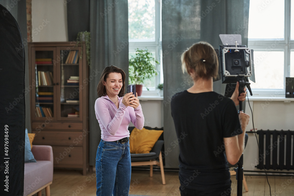 Fototapeta premium Rear view of assistant adjusting strobe light in living room while blogger shooting video on smartphone