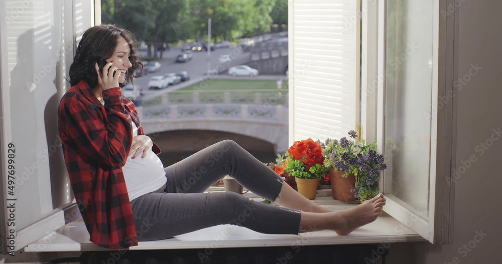 Cute young pregnant woman in casual clothes sit on window sill and talk on smartphone