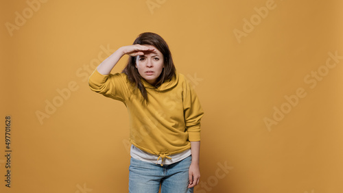 Curious woman looking into the distance far away searching for something with palm over forhead in studio. Young person scouting the horizon seeking for someone over yellow background. photo