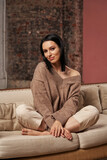 a beautiful woman with black hair in cozy clothes sits on the couch with her legs crossed