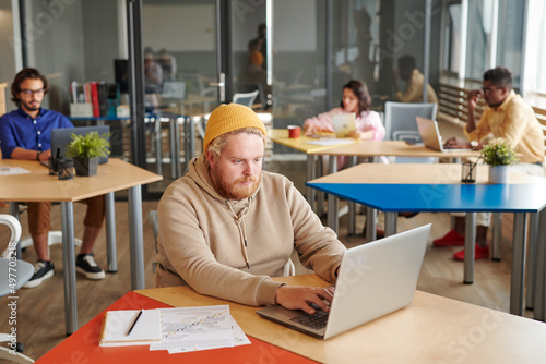 Concentrated young Caucasian manager in yellow hat sitting in coworking space and using laptop while working with online files