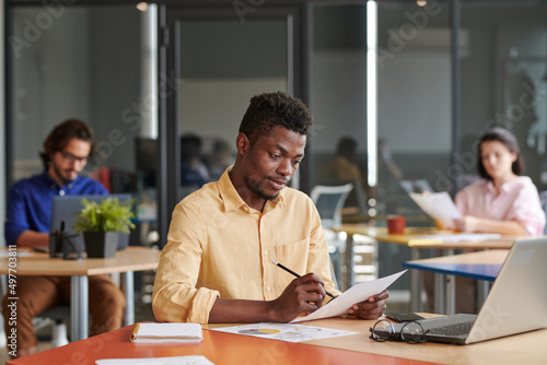 Busy young African-American business expert sitting at table and examining papers in open-space office