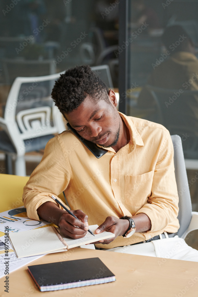 Serious young African-American manager in yellow shirt sitting at table and making notes while calling client Serious young African-American manager in yellow shirt sitting at table and making notes while calling client