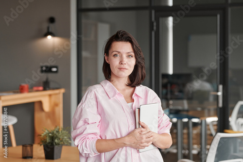 Portrait of content beautiful young Caucasian office woman in pink shirt holding tablet and planner in modern office