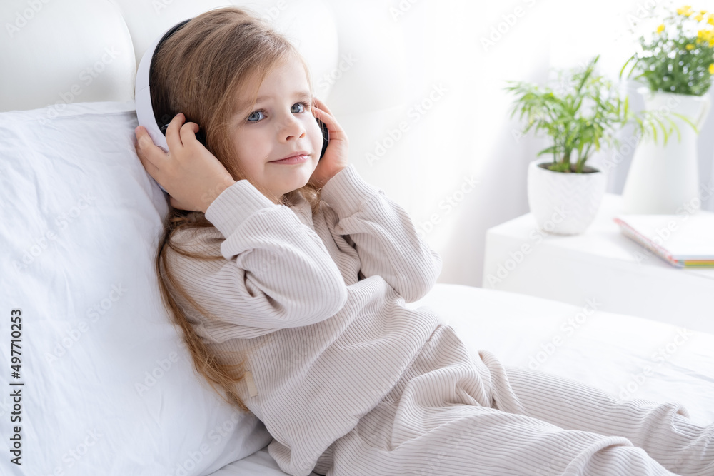 happy little kid caucasian girl in pyjama in wireless headphones listening music on bed at home happy little kid caucasian girl in pyjama in wireless headphones listening music on bed at home
