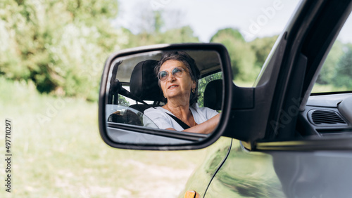 Senior cheerful smiling female driver behind wheel of car looking away, reflection in side rearview mirror outdoors. Caucasian elderly woman driving car, close-up