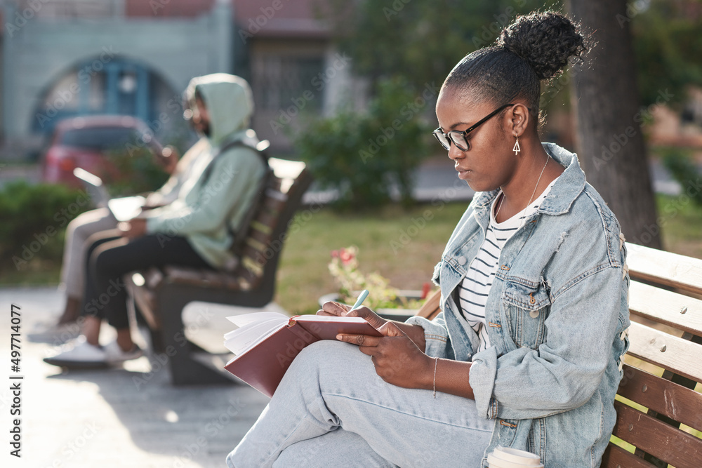 Obraz premium Concentrated young African-American woman in denim outfit and making notes in organizer while working on project in city