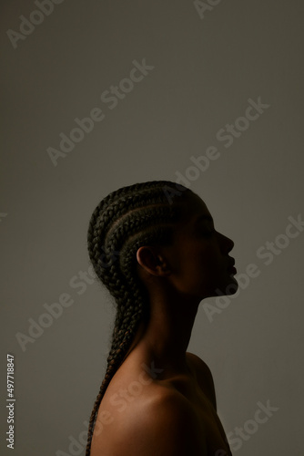 Vertical side portrait of African young woman with braids posing on dark wall.