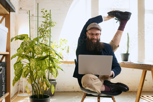 Fototapeta Naklejka Na Ścianę i Meble -  Young bearded man, office clerk having fun, doing yoga on wooden table in modern office at work time with gadgets. Concept of business, healthy lifestyle, sport, hobby