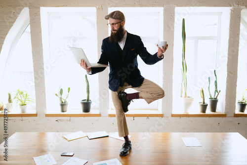 Fototapeta Naklejka Na Ścianę i Meble -  Young bearded man, office clerk having fun, doing yoga on wooden table in modern office at work time with gadgets. Concept of business, healthy lifestyle, sport, hobby
