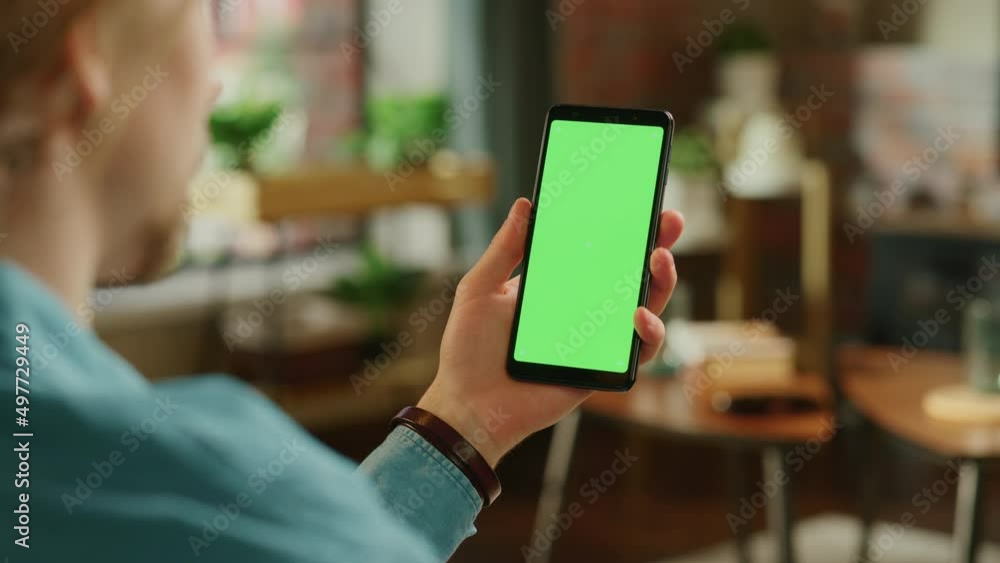 Man Holding a Smartphone with Green Screen Mock Up Display. Male is Resting at Home, Watching Videos and Reading Social Media Posts on Mobile Device. Close Up Over the Shoulder Footage. Man Holding a Smartphone with Green Screen Mock Up Display. Male is Resting at Home, Watching Videos and Reading Social Media Posts on Mobile Device. Close Up Over the Shoulder Footage.