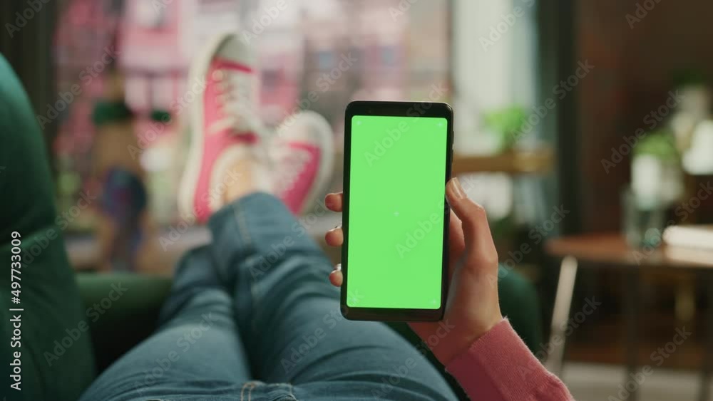 Feminine Hand Holding a Smartphone with Green Screen Mock Up Display. Female is Relaxing on a Couch at Home, Watching Videos and Reading Social Media Posts on Mobile Device. Close Up POV Footage. Feminine Hand Holding a Smartphone with Green Screen Mock Up Display. Female is Relaxing on a Couch at Home, Watching Videos and Reading Social Media Posts on Mobile Device. Close Up POV Footage.
