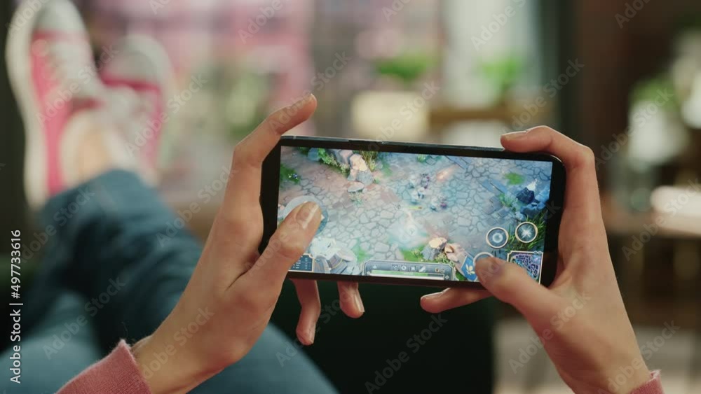Female is Relaxing on a Couch at Home, Playing an Interactive PvP RPG Strategy Video Game on Her Smartphone. Gamer Lies on a Sofa in Living Room. Close Up POV Footage of Mobile Device Screen. Female is Relaxing on a Couch at Home, Playing an Interactive PvP RPG Strategy Video Game on Her Smartphone. Gamer Lies on a Sofa in Living Room. Close Up POV Footage of Mobile Device Screen.