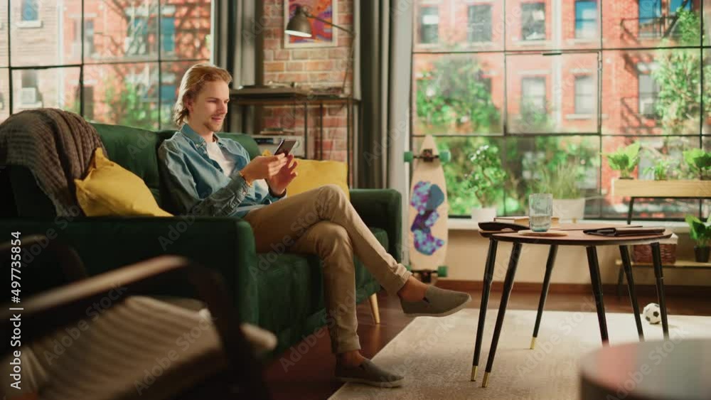 Young Handsome Adult Man Sitting on a Couch in Loft Apartment, Relaxing and Using Smartphone. Creative Male Checking Social Media, Chatting with Friends, Browsing Internet. City View from Big Window. Young Handsome Adult Man Sitting on a Couch in Loft Apartment, Relaxing and Using Smartphone. Creative Male Checking Social Media, Chatting with Friends, Browsing Internet. City View from Big Window.