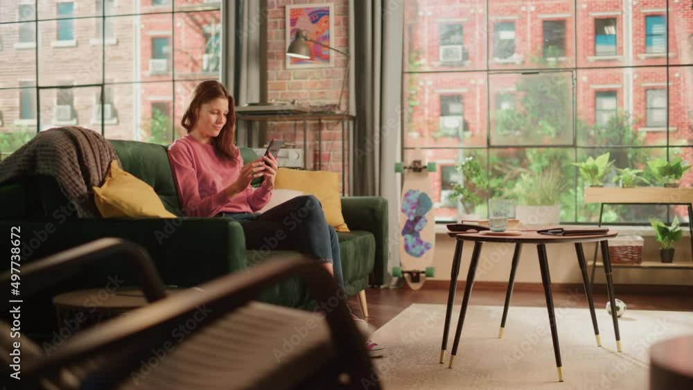 Young Beautiful Female Sitting on a Couch in Living Room, Resting and Using Smartphone. Creative Girl Checking Social Media, Chatting with Friends, Browsing Internet. City View from Big Window. Young Beautiful Female Sitting on a Couch in Living Room, Resting and Using Smartphone. Creative Girl Checking Social Media, Chatting with Friends, Browsing Internet. City View from Big Window.