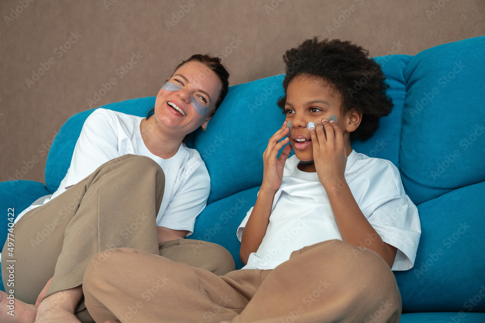 Happy Caucasian mother and an African American daughter are sitting at home on the couch with patches on their faces,having fun.Diverse people.Time together.Mother's day.Selective focus. Happy Caucasian mother and an African American daughter are sitting at home on the couch with patches on their faces,having fun.Diverse people.Time together.Mother's day.Selective focus.