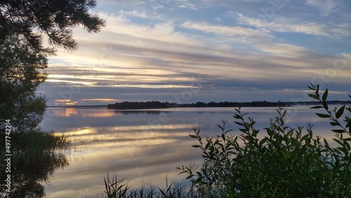 On a summer morning  the sun rises over the lake and illuminates the clouds. The sky and clouds are reflected in the calm water. Reeds grow in the water. Branches of trees lean over the water