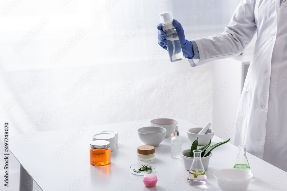 partial view of laboratory assistant in latex glove holding bottle with cosmetic product near ingredients in lab. partial view of laboratory assistant in latex glove holding bottle with cosmetic product near ingredients in lab.