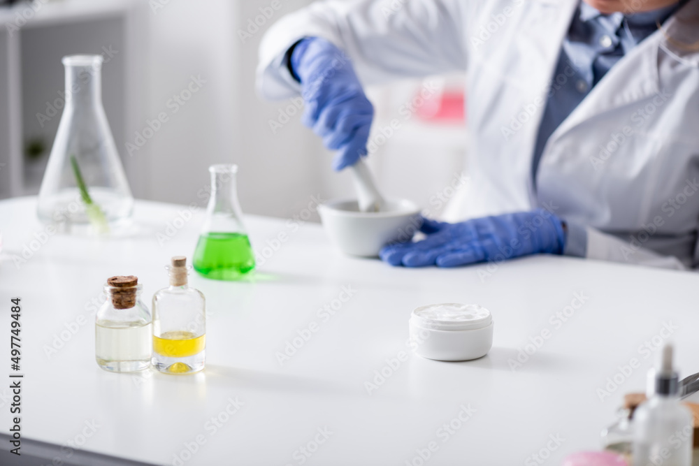 flasks and bottles near blurred laboratory assistant in latex gloves working in lab. flasks and bottles near blurred laboratory assistant in latex gloves working in lab.