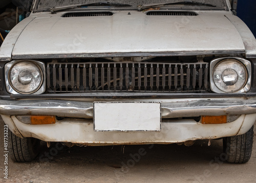 Picture of an old white classic car parked in the garage. Take a photo of the front of the car looking shabby.The hood and the metal fenders are rusted.