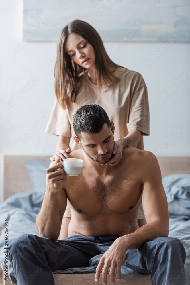 Fototapeta premium Blurred woman in t-shirt touching muscular man holding cup on bed.