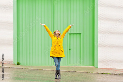 Horizontal front view of woman raising arms in a yellow raincoat standing on the street. Full length body of woman under the rain isolated on green wall. People isolated in background concept.