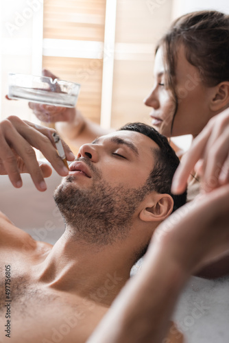 Sexy man holding cigarette near blurred girlfriend with ashtray in bathtub.
