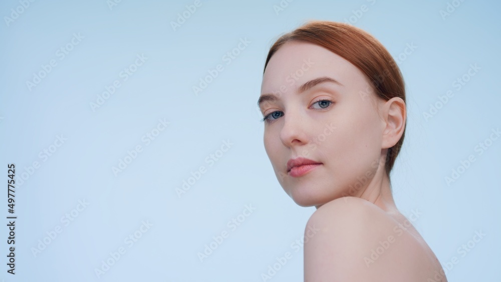 Low-angle beauty portrait of young redhead woman who looks at camera over shoulder against blue background Low-angle beauty portrait of young redhead woman who looks at camera over shoulder against blue background