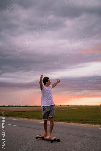 Freedom and active happy people concept lifestyle with young boy moving on skateboard lonboard on asphalt road outstretching arms agains a colorful amazing sunset background