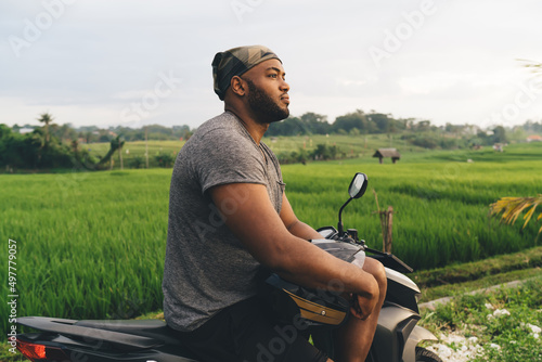 Contemplative male tourist with helmet resting at motorbike thinking about driving trip during summer vacations in Indonesia, African American hipster guy in bandanna feeling pondering on rent details #497779057