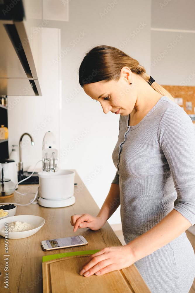 Pretty woman using smartphone, reading recipe online, looking at phone screen, healthy home food, in modern kitchen Pretty woman using smartphone, reading recipe online, looking at phone screen, healthy home food, in modern kitchen
