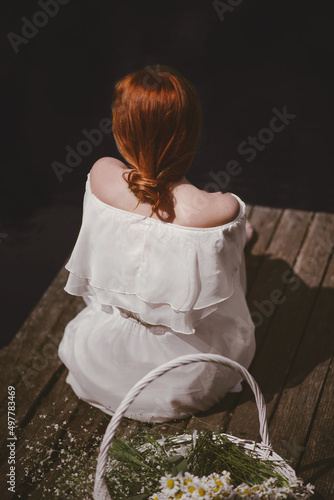 beautiful young girl with red hair gathered in a bundle in a white dress sitting on a wooden pier next to her stands a white straw basket with field rum rural scene on a sunny day rear view  photo