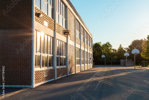 The exterior of the school building and school yard with a basketball court on a sunny evening. The sun ireflecting in the windows