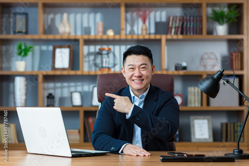 Happy asian businessman showing finger aside sitting in office, smiling and looking at camera © Liubomir