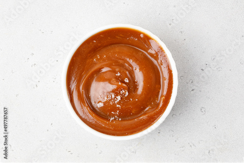 Salted caramel sauce in a white saucer on grey background. Curl of caramel with sea salt pieces. Top view.