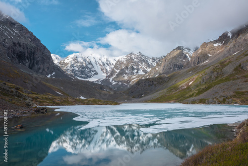 Atmospheric landscape with frozen alpine lake and high snow mountains. Ice floats on transparent water surface of mountain lake. Awesome scenery with large snowy mountains reflection in clear lake.