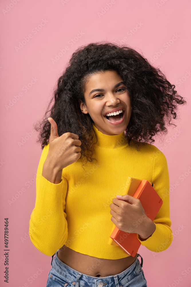 Portrait of young happy African American student holding books showing thumb up looking at camera isolated on pink background. Education concept Portrait of young happy African American student holding books showing thumb up looking at camera isolated on pink background. Education concept