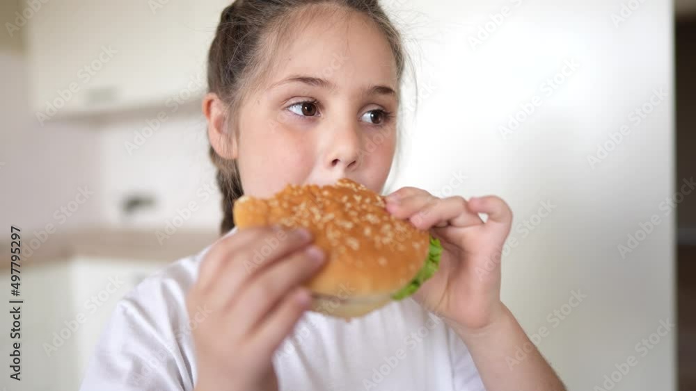 little girl eating a hamburger. unhealthy fast food proper nutrition concept. child greedily with pleasure bites a big burger in the kitchen at home. kid eats fast lifestyle food close-up little girl eating a hamburger. unhealthy fast food proper nutrition concept. child greedily with pleasure bites a big burger in the kitchen at home. kid eats fast lifestyle food close-up