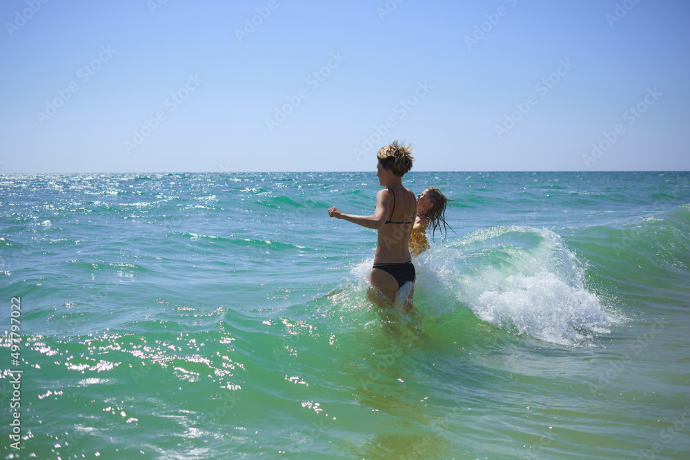Summer happy family of six years blonde child playing and jumping water waves embracing woman mother in sea shore beach Summer happy family of six years blonde child playing and jumping water waves embracing woman mother in sea shore beach